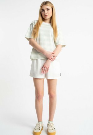 Młoda kobieta z długimi blond włosami stoi, ubrana w lekką paskowaną koszulkę, białe szorty i beżowe buty sportowe z gumową podeszwą na białym tle.