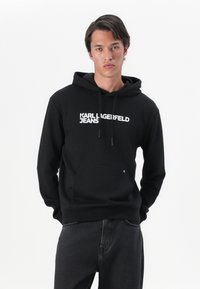 Čierna mikina s kapucňou vyrobená z bavlnenej zmesi, s predným klokankovým vreckom, šnúrkami a bielym logom "KARL LAGERFELD JEANS". Minimalistický dizajn.