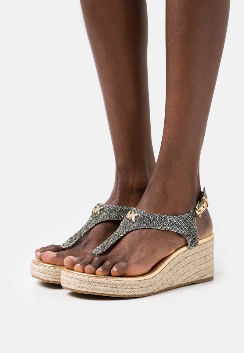 Gros plan sur des pieds portant des sandales compensées pailletées avec le logo MK sur les brides et une semelle en corde de jute, posés sur un fond blanc.