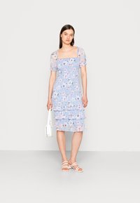 Vestido floral com mangas bufantes em azul claro, apresentando babados em camadas e decote quadrado. Combinado com sandálias brancas e uma bolsa.