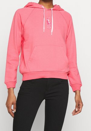 Personne portant un sweat à capuche rose avec des cordons blancs et un petit logo de cheval brodé, associé à un pantalon noir, raccourci au niveau du torse.