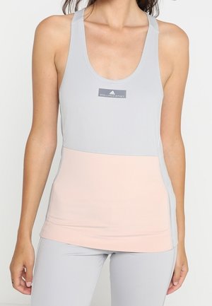 Kobieta ubrana w bezrękawnik sportowy w jasnoszarym i różowym kolorze z logo na piersi oraz dopasowane jasnoszare legginsy.