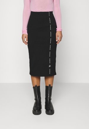 Zwarte midi-rok met een aansluitende pasvorm, voorzien van witte logotekst verticaal langs de zijkant. Gecombineerd met zwarte laarzen tot aan de knie.