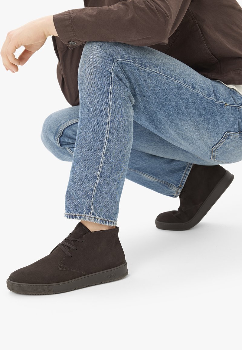 Personne portant un jean bleu et des chaussures en daim marron foncé à lacets, accroupie avec un genou plié sur un fond blanc.
