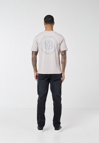 Bărbat cu brațe tatuate, purtând un tricou gri deschis cu text și numere albastre circulare, blugi negri și pantofi negri cu talpă albă, în picioare.