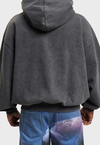 Felpa grigia oversize con finitura testurizzata e cappuccio, abbinata a pantaloni in denim blu con motivo astratto colorato e accento con logo.