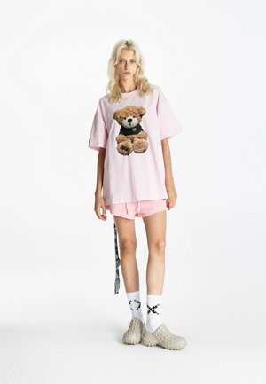 Joven mujer con camiseta rosa oversize con dibujo de oso de peluche, pantalones cortos rosas, calcetines blancos con diseño negro y zapatos beige texturizados sin cordones.
