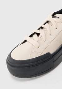 Off-white nubuk tenisice s debelim crnim gumiranim potplatom i odgovarajućim off-white vezicama, prikazane s prednje i bočne strane na bijeloj pozadini.