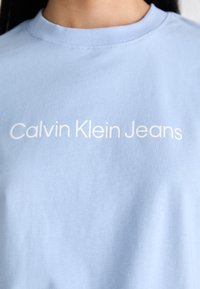 Světle modré bavlněné tričko s kulatým výstřihem a textem "Calvin Klein Jeans" v bílé barvě, umístěným ve středu hrudi.