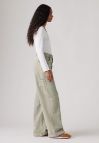 Γυναίκα με μακριά σγουρά μαλλιά, φορώντας λευκό μπλουζάκι με μακριά μανίκια, ανοιχτοπράσινο παντελόνι με φαρδιά πόδια και πορτοκαλί αθλητικά, στάζει πλευρικά σε απλό φόντο.