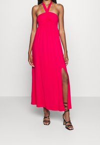Maxikleid in kräftigem Pink, aus weichem Stoff, mit Halterneck, gerafftem Oberteil und einem seitlichen Schlitz. Kombiniert mit schwarzen Riemchensandalen.