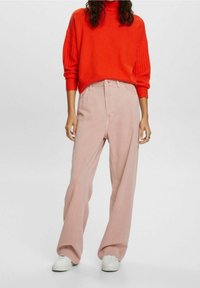Roter gerippter Strickpullover mit hohem Halsausschnitt, kombiniert mit hellrosa weit geschnittenen Hosen und weißen Sneakern, was ein lässiges Outfit ergibt.