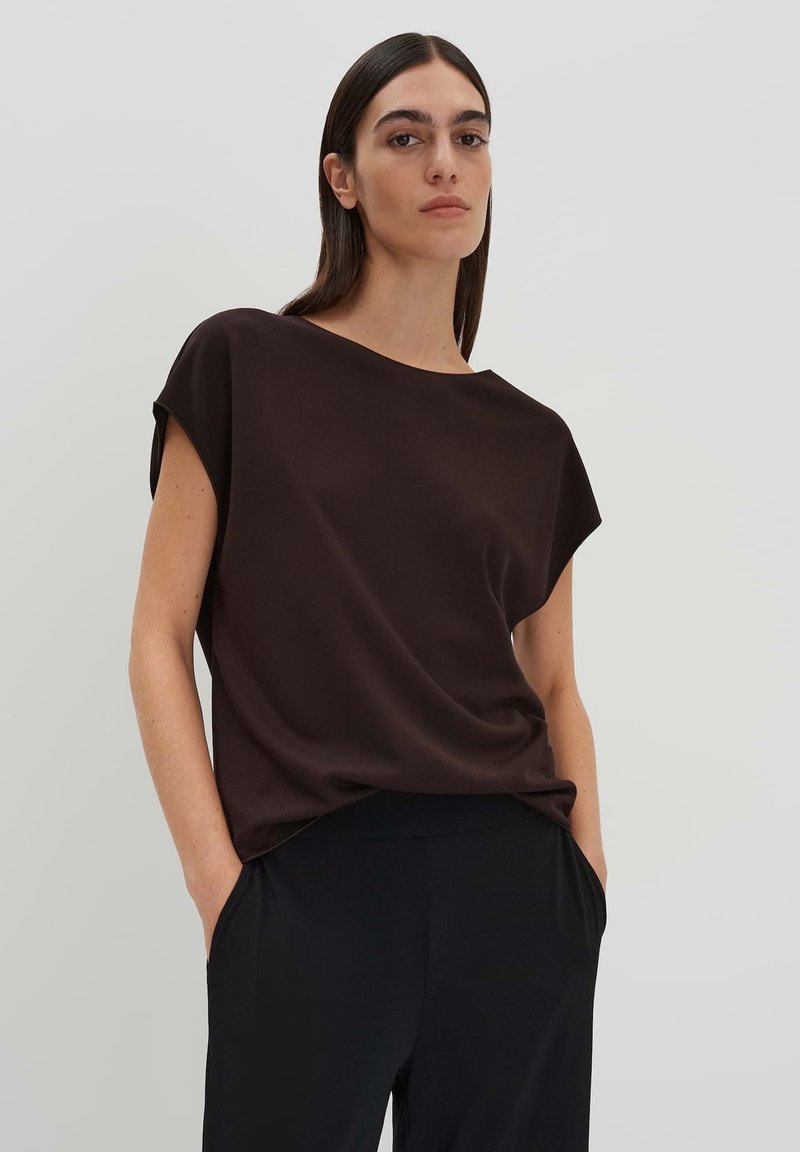Top marrone a maniche corte con vestibilità rilassata e texture liscia, abbinato a pantaloni neri. Il top presenta un orlo asimmetrico.