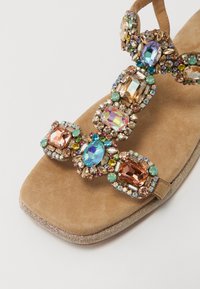 Flat sandal med en brun semsket skinnfotbed, pyntet med fargerike fasetterte edelstener og krystalldetaljer, med en firkantet tå og en subtil glitrende stropp.