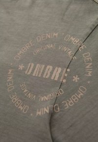 Šedá látka s měkkou texturou, na které se nachází kruhový, světle béžový potisk s nápisem "OMBRE DENIM ORIGINAL VINTAGE" a dekoračními prvky.