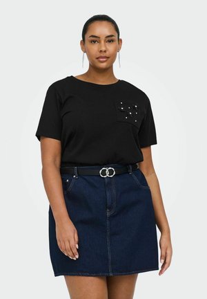 Kvinne iført svart kortermet topp med små blomsterapplikasjoner på lommen, et svart belte med sølvspenne og et mørkeblått denimskjørt.