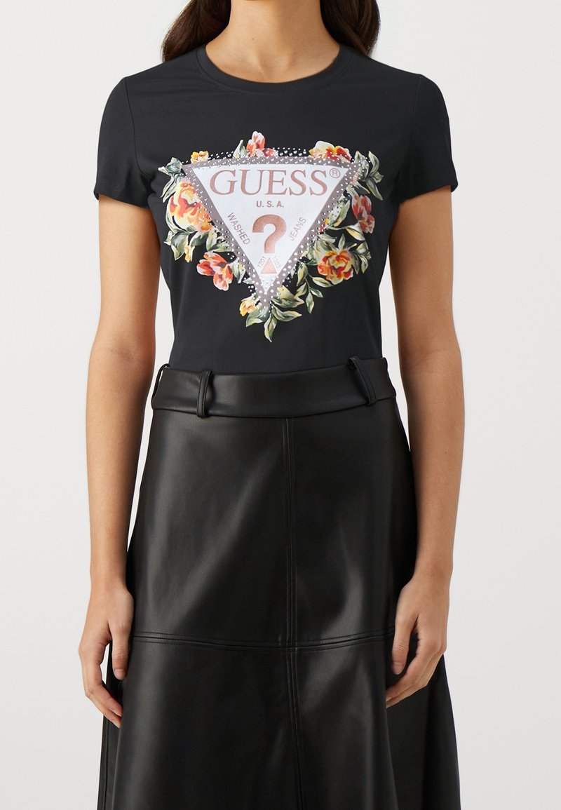 Sort kortærmet t-shirt med et blomstertriangel grafik med "GUESS" tekst og glitrende accenter, parret med en sort lædernederdel.