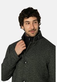 Grauer Hahnentrittmantel mit hohem Kragen, schwarzem Innenfutter und Knopfverschluss. Die Textur ist gewebt und leicht strukturiert.