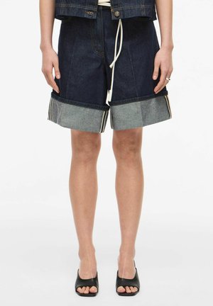 Personne portant un short en jean foncé retroussé, des talons ouverts noirs et une veste en jean foncé assortie avec un cordon blanc.