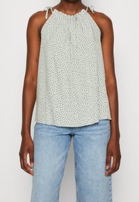 Lichtgroene mouwloze top met een klein wit bloemenpatroon, geplooide halslijn en strikbandjes. Gecombineerd met lichtblauwe jeans.