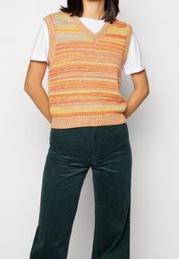 Stickad ärmlös tröja i klara orange, rosa och gula ränder med V-ringning, kombinerad med mörkgröna kordbyxor med vertikala ribbor.