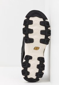 Svart atletisk sko med en texturerad ovandel, en tjock vit mellansula och en robust svart yttersula med djupa mönster och en guld-logotyp som accent.