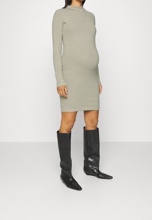 Vrouw draagt een nauwsluitende, lange mouw, beige en zwart gestreepte jurk tot op de knie, gecombineerd met zwarte knielange leren laarzen.