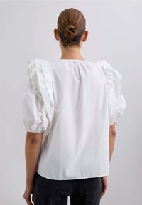 Weiße Bluse mit puffigen Ärmeln und Spitzenbesatz sowie rundem Ausschnitt. Glatte Textur, Rückansicht.