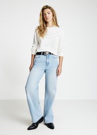 Weißes langärmliges Sweatshirt mit strukturiertem Detail, hellblaue Jeans mit weitem Bein, schwarzer Gürtel mit silberner Schnalle und schwarze Stiefel mit spitzer Kappe.