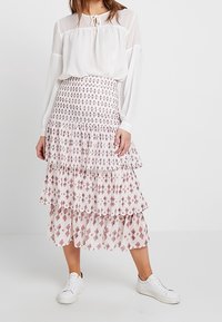 Valkoinen pusero, jossa on pitkät hihat ja solmu kaulassa, yhdistettynä kerrokselliseen midi-hameeseen, jossa on pinkki geometrinen kuvio. Asusteena valkoiset lenkkarit.