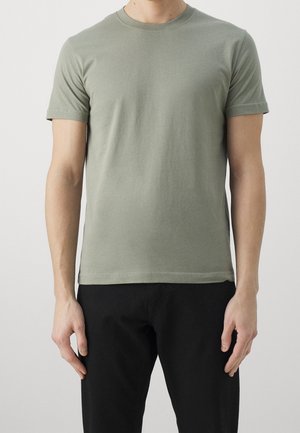 Mężczyzna ubrany w gładki, jasnooliwkowy t-shirt z krótkim rękawem i czarne spodnie, stojący na gładkim, jasnym tle.