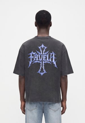 Άνδρας που φοράει σκούρο φανταχτερό T-shirt με μπλε στυλιζαρισμένο σταυρό και τη λέξη "FAVELA" στην πλάτη, συνδυασμένο με ανοιχτό μπλε τζιν.