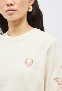 Γυναίκα που φοράει χρυσούς κρίκους στα αυτιά και ένα κρεμ φούτερ με μικρό ροζ κεντημένο πέταλο αλόγου στο στήθος και κεντήματα με λουλούδια στο μανίκι.