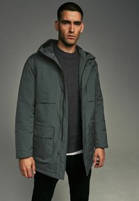 Chaqueta acolchada gris con capucha, que cuenta con múltiples bolsillos frontales y cierre de cremallera. Lleva encima un suéter de punto oscuro y pantalones negros.