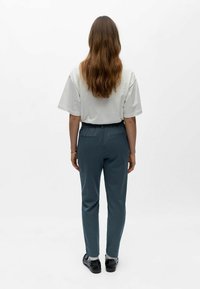 Femme aux longs cheveux ondulés portant une chemise blanche à manches courtes, un pantalon bleu foncé et des chaussures noires, se tenant de dos sur fond blanc.