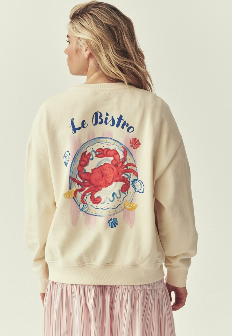 Femme aux cheveux blonds porte un sweat-shirt beige avec un crabe rouge sur une assiette avec quartiers de citron et coquillages, texte « Le Bistro » au-dessus.