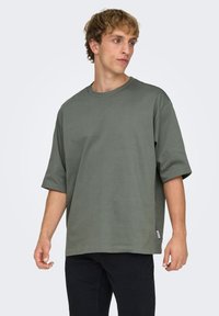 T-shirt surdimensionné vert olive en coton doux, avec un col rond et des manches courtes, associé à un pantalon en jean noir.