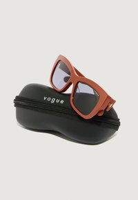 Ochelari de soare roșii cu rame pătrate așezați pe un toc negru cu fermoar, prezentând cuvântul "vogue" în text alb.