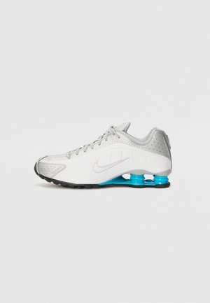 White and gray Nike running shoe with black sole and blue spring-like cushioning in the heel on a plain white background.