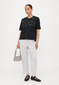 T-shirt noir avec des embellissements en argent décoratifs, associé à un pantalon gris clair et des chaussures noires. Le modèle tient un petit sac à main texturé.