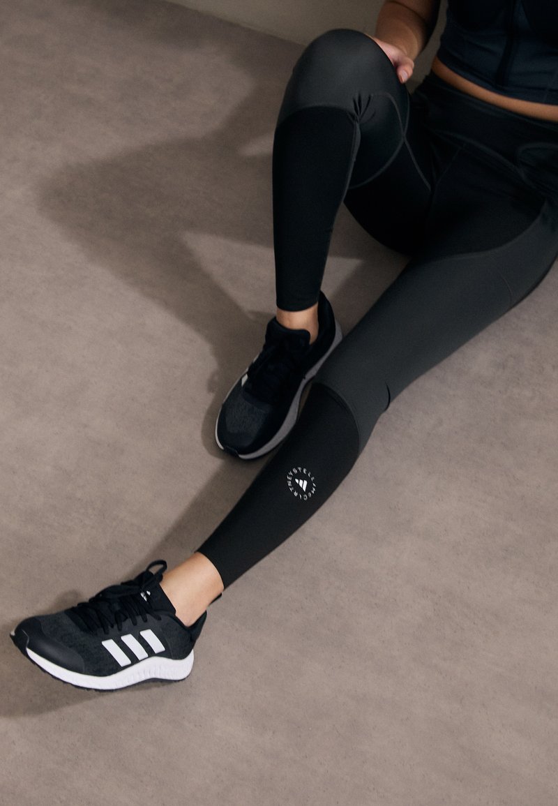 Sorte treningsleggings med en logo på leggen, sammen med svarte og grå løpesko med tre hvite striper og et teksturert design.