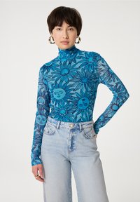 Blauwe bloeiende mesh coltrui met zonmotieven, gecombineerd met hoog-getailleerde lichte spijkerbroek. Gladde textuur, lange mouwen, getailleerd ontwerp.