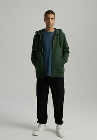 Zielona bluza z zamkiem i kieszeniami z przodu, nałożona na niebieską koszulkę, zestawiona z czarnymi spodniami cargo i białymi trampkami. Gładkie tło.