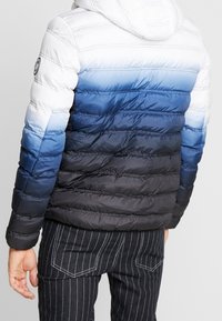 Heren puffer jacket met een verloop van wit naar marineblauw. Kenmerkt zich door horizontale quilting en een capuchon. Gekocht bij gestreepte broeken.
