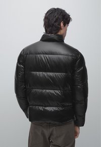 Man draagt een zwarte glanzende donsjack, gezicht van de kijker afgewend, gecombineerd met donkergrijze broek tegen een effen lichtgrijze achtergrond.