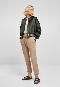 Veste bomber noir en satin avec des poignets à rayures blanches, joggers beiges avec cordon de serrage et sandales noires. Le mannequin se tient les mains dans les poches.
