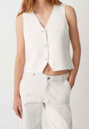 Femme portant un gilet blanc sans manches boutonné et un pantalon blanc taille haute, avec une main dans la poche, devant un fond uni.