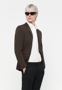 Blazer personalizat maro peste o cămașă albă și pantaloni negri. Modelul poartă ochelari de soare negri. Material neted cu croială structurată și linii curate.