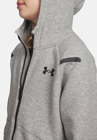 Grijze zip-up hoodie met een zachte textuur, voorzien van een zwarte ritssluiting, zwart logo en zwarte accenten op de schouders. Met capuchon.