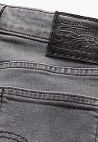 Jeans de mezclilla gris con una superficie texturizada, que presentan una etiqueta de cintura de cuero negro y detalles de costura en marrón.